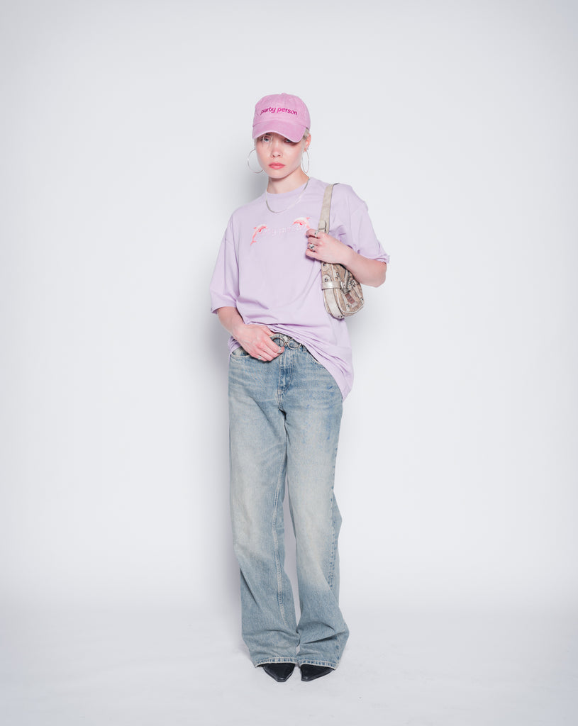 Model wearing washed pink dad cap with party person embroidery and lilac oversize t-shirt with party person print.